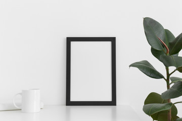 Black frame mockup with a mug, book and ficus on a white table. Portrait orientation.