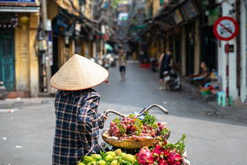 The street vendor with bike loaded of tropical fruits in old town street in Hanoi, old houses and street activites on background