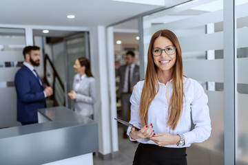 Charming Caucasian businesswoman dressed in shirt and skirt standing on hallway and holding tablet. In background her colleagues standing and chatting. - obrazy, fototapety, plakaty