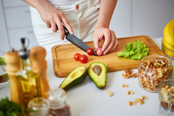 Young happy woman cutting tomatoes for making salad in the beautiful kitchen with green fresh ingredients indoors. Healthy food and Dieting concept. Loosing Weight