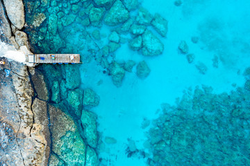 An aerial view of the beautiful Mediterranean Sea, with a wooden pier and a rocky shore, where you can see the textured underwater corals and the clean turquoise water of Protaras, Cyprus