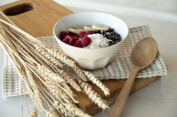 Delicious healthy breakfast of cottage cheese with berries. A white plate with cottage cheese and berries stands on a wooden board on a white table, next to it are wheat ears. Diet.