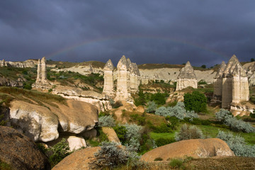Love valley in Goreme village, Turkey. Rural landscape. Beautiful sunny light on stone houses with rain, rainbow and blue dark sky background. Cappadocia