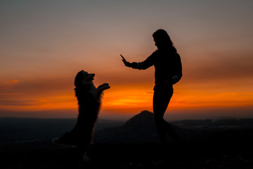 Silhouette of young woman with his dog doing trick at dawn with mountain on background.