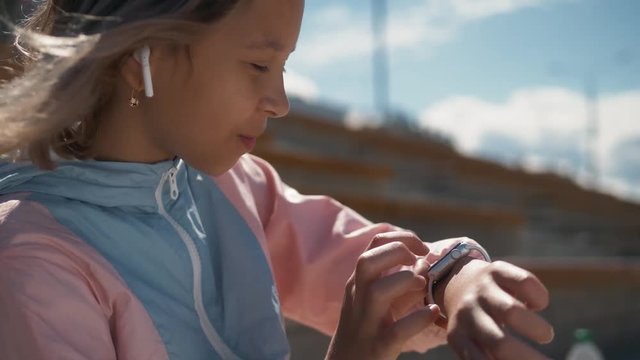 Kid Using Smartwatch or Smart Watch in park sitting on grass, Child in garden with airpods headphones dictate audio message with voice recognition