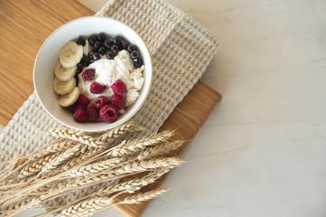 Delicious healthy breakfast of cottage cheese with berries. A white plate with cottage cheese and berries stands on a wooden board on a white table, next to it are wheat ears. Diet.