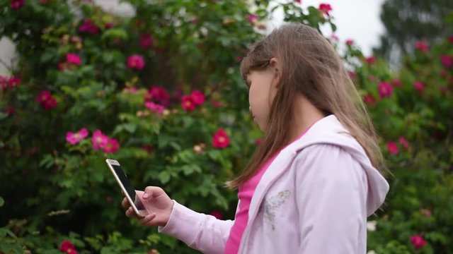 European girl with blonde hair taking pictures of herself on her smartphone. Girl takes a selfie on the phone in the garden on a background of blooming roses