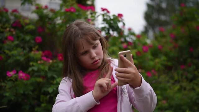 European girl with blonde hair taking pictures of herself on her smartphone. Girl takes a selfie on the phone in the garden on a background of blooming roses