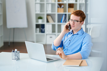 Fototapeta premium Young serious manager in blue shirt talking by smartphone by desk