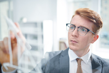 Young elegant student in suit and eyeglasses looking at transparent board