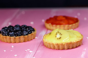 Freshly baked tartlets with strawberry, banana, blueberry fruit filling on a pink wooden background