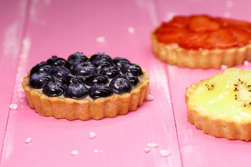Freshly baked tartlets with strawberry, banana, blueberry fruit filling on a pink wooden background