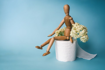 Wooden figure sit on a roll of toilet paper and flower of buldenege on a blue background. Toilet...