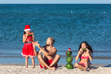 Merry family celebrates summer Christmas. father and two daughters eating watermelon on beach.