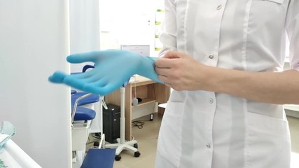 A female doctor surgeon in white uniform puts on blue gloves before surgery. Gynecological cabinet with chair and other medical equipment on background. Concept of hygiene, treatment, medicine.
