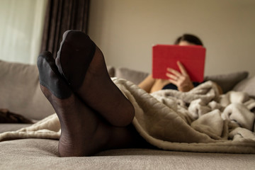 A portrait of crossed feet in pantyhose of a woman relaxing using her tablet in the couch under a blanket at home to relax.