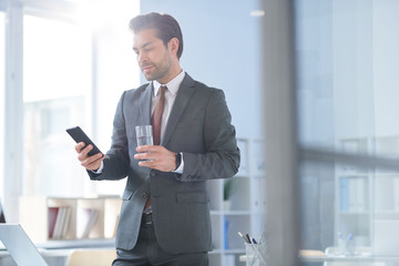 Fototapeta premium Young confident agent with glass of water scrolling in smartphone