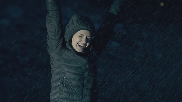pretty young woman in green jacket with hood smiles and enjoys heavy rain at night close view slow motion