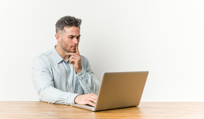 Young handsome man working with his laptop looking sideways with doubtful and skeptical expression.