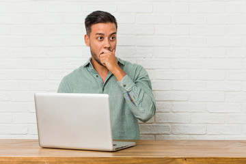 Young filipino man sitting working with his laptop thoughtful looking to a copy space covering mouth with hand.