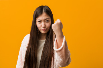 Young chinese woman in pajama showing fist to camera, aggressive facial expression.