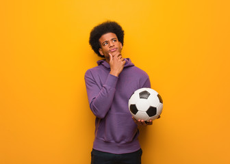 Young african american sport man holding a soccer ball doubting and confused