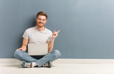 Young redhead student man sitting on the floor pointing to the side with finger. He is holding a...
