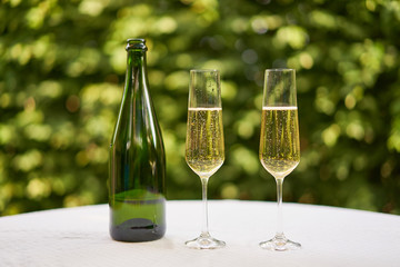 Two wine glasses flute type with sparkling wine or champagne and traditional design champagne bottle on the table with white tablecloth in the romantic garden restaurant ready to celebrate or cheers.