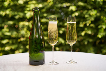 Close up Picture on two wine glasses flute type with sparkling wine or champagne and traditional design champagne bottle on the table with white tablecloth in the romantic garden restaurant, cheers.