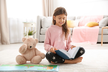 Cute little girl with teddy bear reading book on floor at home