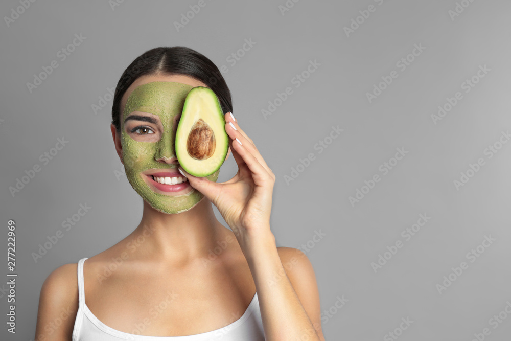 Sticker Young woman with clay mask on her face holding avocado against grey background, space for text. Skin care