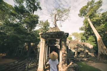One tourist visiting Angkor ruins amid jungle, Angkor Wat temple complex, travel destination Cambodia. Woman with traditional hat, rear view.