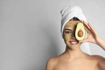 Young woman with clay mask on her face holding avocado against light background, space for text....