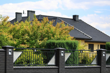 Murowane ogrodzenie z cegły i stali, dach budynku i zielone drzewa.	