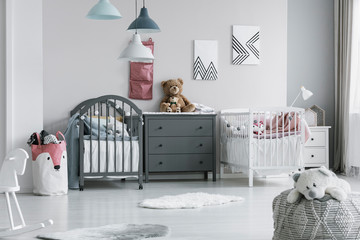Brown teddy bear on grey wooden commode between two cribs in fashionable bedroom for twins