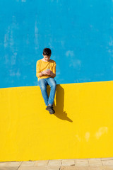 Fototapeta na wymiar Front view of a young boy wearing casual clothes sitting on a yellow fence against a blue wall while using a smartphone