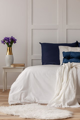 Purple flowers in vase and books on wooden nightstand table next to white bed with navy blue pillows