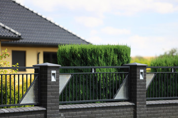Fototapeta premium Murowane ogrodzenie z cegły i stali, dach budynku i zielone drzewa.