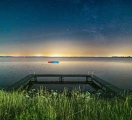 Milky way shot above the water Harbour of Ouddorp, the Netherlands with light pollution, grass and a wooden pier in the forground