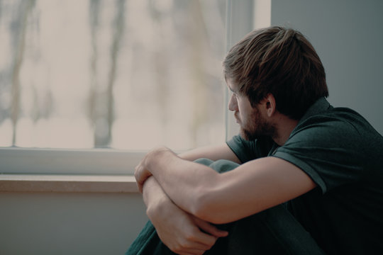 Sad Young Man Sitting On The Floor Looking Through The Window