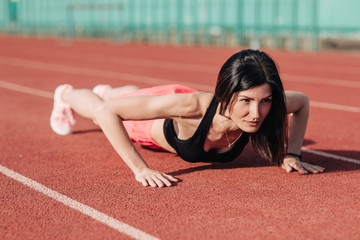 front view of young slim attractive brunette woman in pink shorts and black top doing plank exercise at outdoor stadium, core training and fitness concept, sun flare