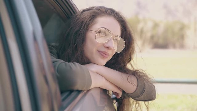 pretty dreaming woman in fashionable glasses looks out of standing automobile window at spring meadow