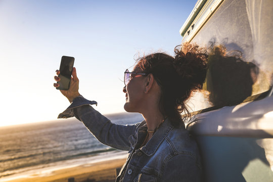 Happy woman taking selfie with smartphone leaning against a vintage van near a beautiful ocean beach. Travel blogger making social video story for blog. New trends technology and communication concept