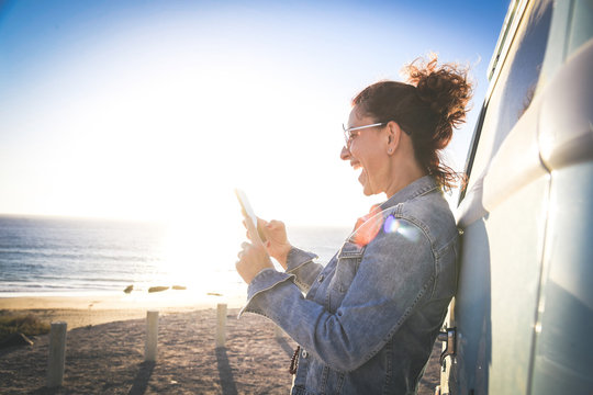 Happy woman laughing watching social media video online. Young girl communicate with remote friends with smartphone leaning against a van near a beach. New trends technology and communication concept - Powered by Adobe