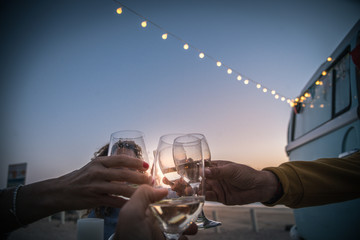 Happy friends cheering and drinking wine during camping vacation at the beach with vintage van. People having fun at weekend summer evening with camper. Youth lifestyle togetherness and party concept