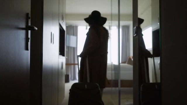Back view woman opening door and entering in hotel apartment. Follow faceless female in hat with suitcase moving room, coming to full length window. Concept comfortable vacation