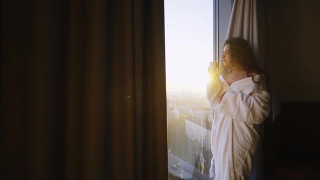 Young woman relax in modern hotel apartment. Side view girl in white bathrobe standing near full length window in sun shining, enjoying morning coffee and cityscape through window