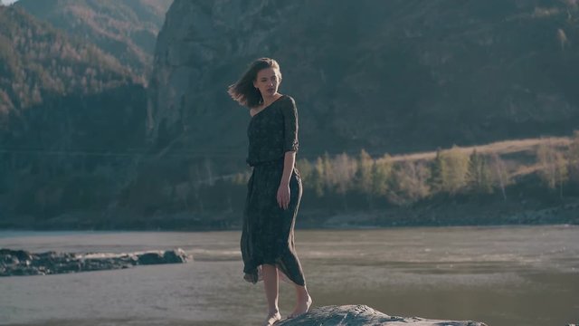 young graceful woman in dark dress stands on rock near wide river against mountains on windy day