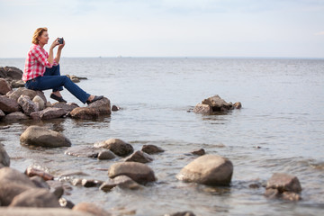 Caucasian woman sitting on stone near the water. Making pictures with cellphone, calm sea