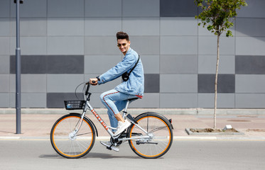 Stylish guy dressed in trendy jeans clothes rides bicycle in the street against the background of a gray building